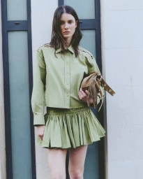 Model wearing a sage green button-up shirt and matching pleated skirt holding a fringed brown bag against a stone exterior wall