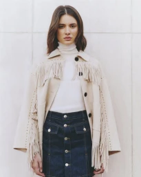 Model wearing a fringed beige jacket and dark denim skirt standing against a light panelled wall