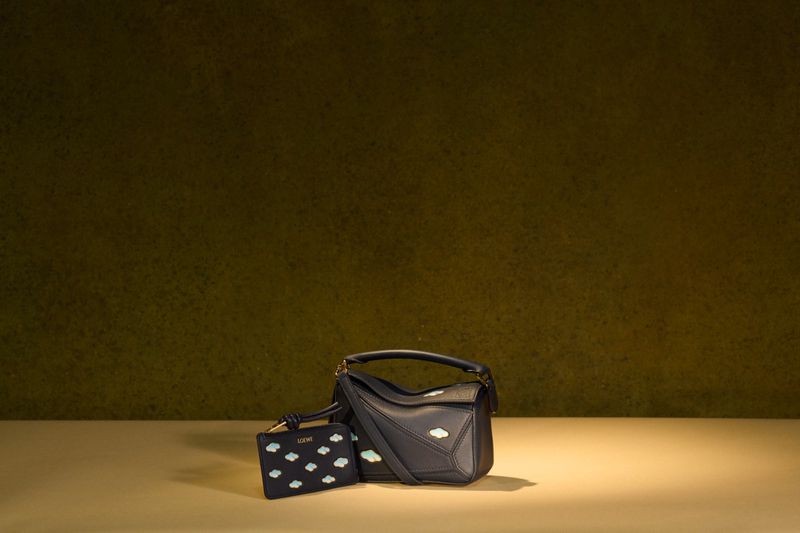 Dark blue leather Loewe Puzzle bag and cardholder with cloud patterns on a tan surface against a textured green background in a studio shot.