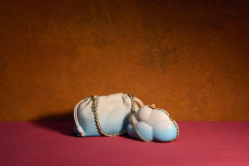 Two ombre handbags, one a cloud shape, sit on a magenta surface against a textured umber background, lit by soft studio lighting.