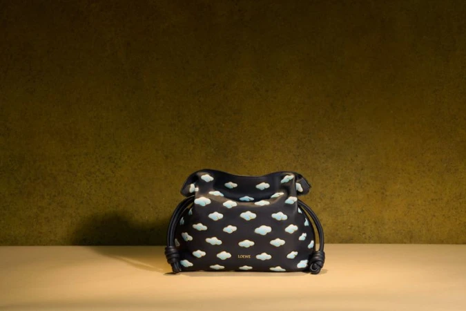 Close-up studio shot of a Loewe black drawstring pouch with blue cloud pattern, set against a muted gold background. Product photography.