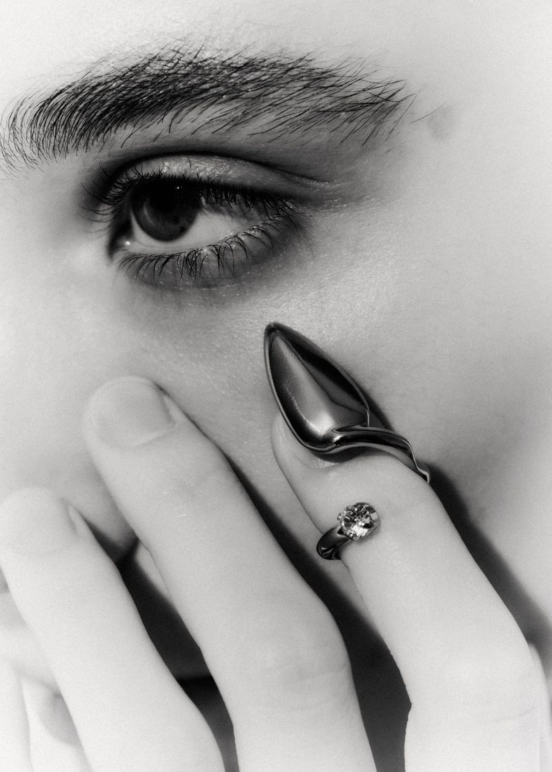 Close-up of a person wearing a metallic claw ring near their eye, photographed in black and white.