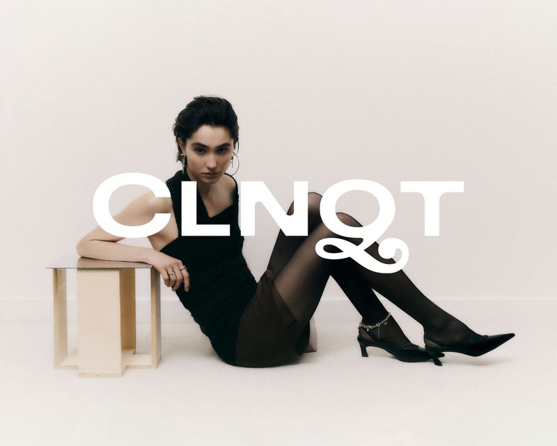 Young woman in a black dress poses in front of a white background sitting on a small table with a reflective surface in a studio shot.