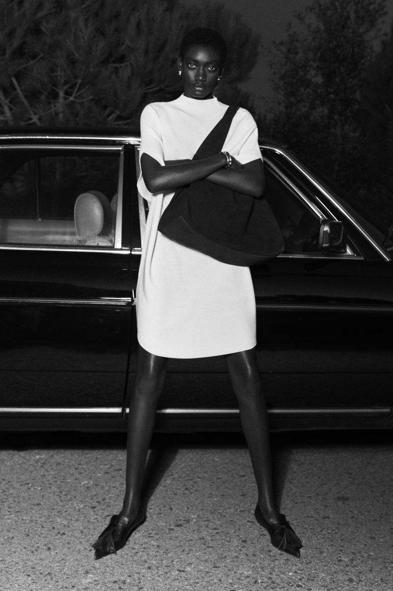 Monochrome portrait of an adult Black woman in a white dress and black bag, standing in front of a vintage car with trees in the background.