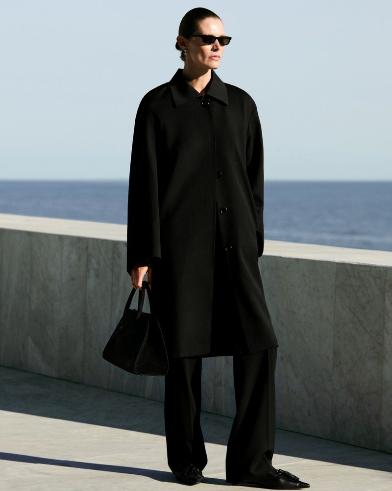 Model in black coat holding a handbag standing against a stone wall with ocean background