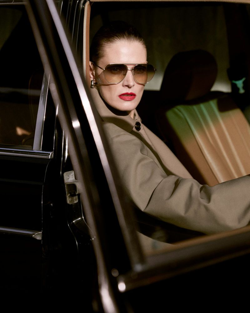 A brunette adult female wears sunglasses and a jacket while sitting in a car with the door open, posing for a fashion-style portrait.