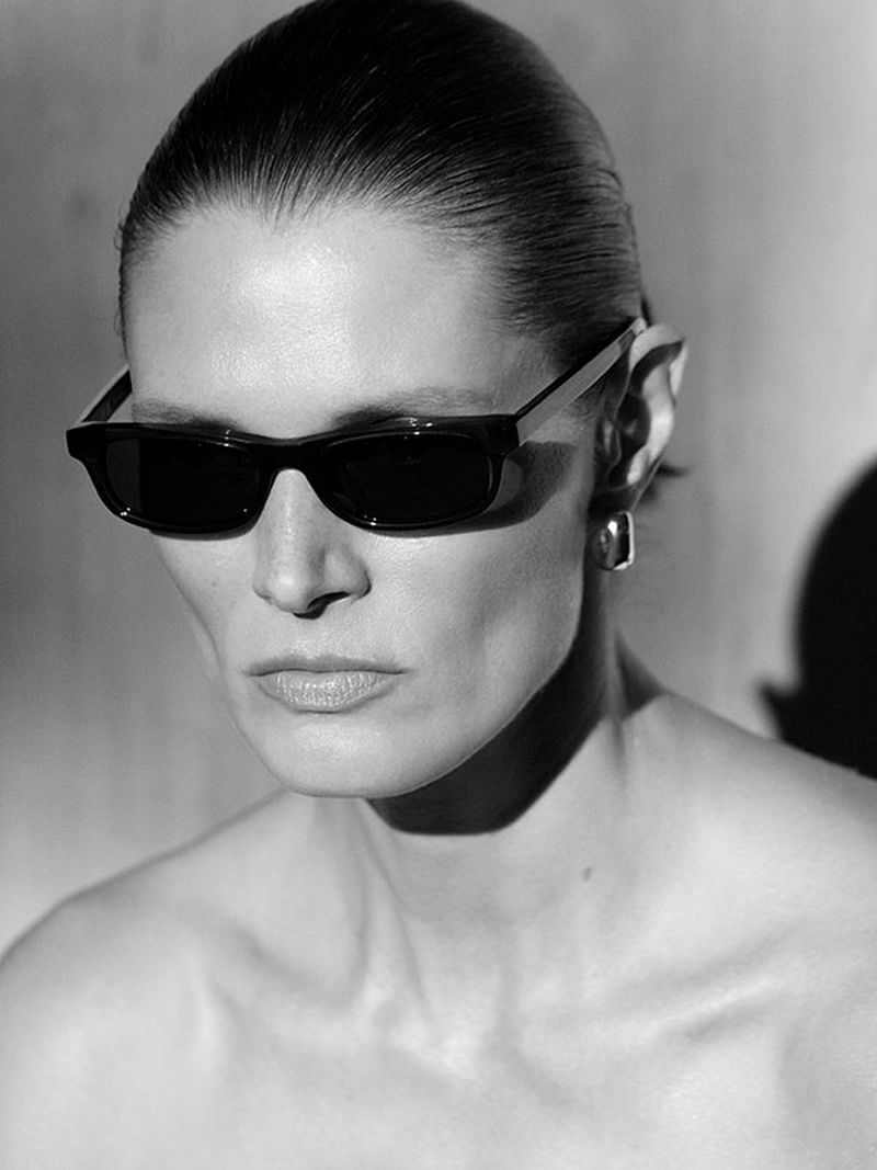 Monochrome portrait of a woman with sunglasses and bare shoulders against a plain backdrop, captured in high-contrast lighting.