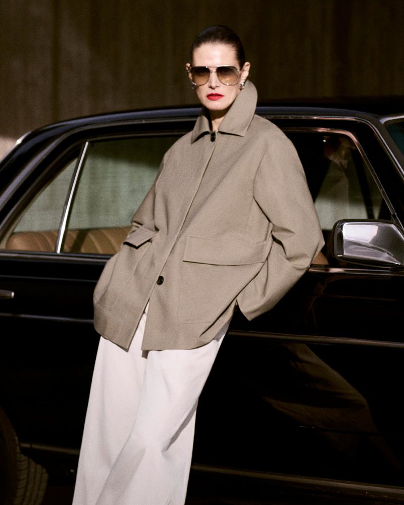 Stylish woman in sunglasses and beige jacket poses by black car in an outdoor setting, showcasing fashion and automotive aesthetics.