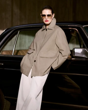 Stylish woman in sunglasses and beige jacket poses by black car in an outdoor setting, showcasing fashion and automotive aesthetics.