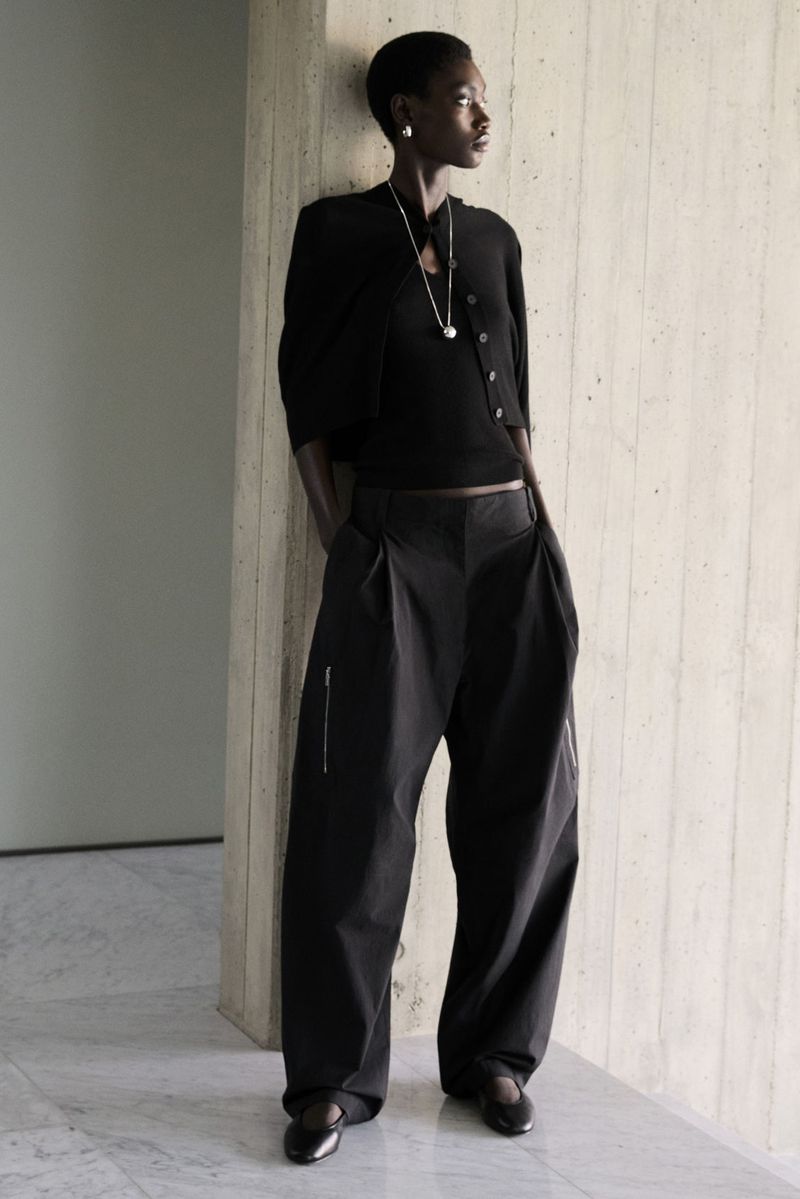 Fashionable adult female with short hair poses in black cardigan and trousers against a textured wall in a full-length studio shot.