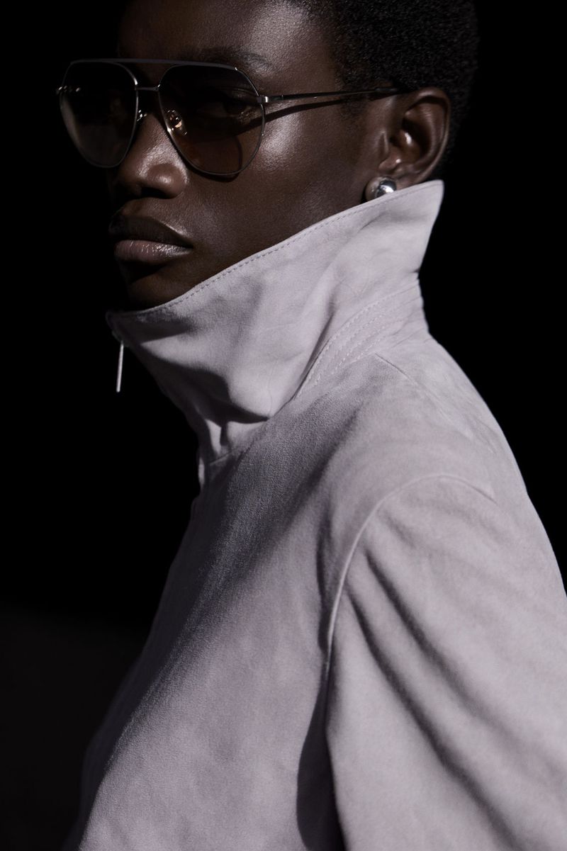 Close-up of a dark-skinned woman wearing aviator sunglasses and a high-collared jacket, against a dark background in studio lighting.