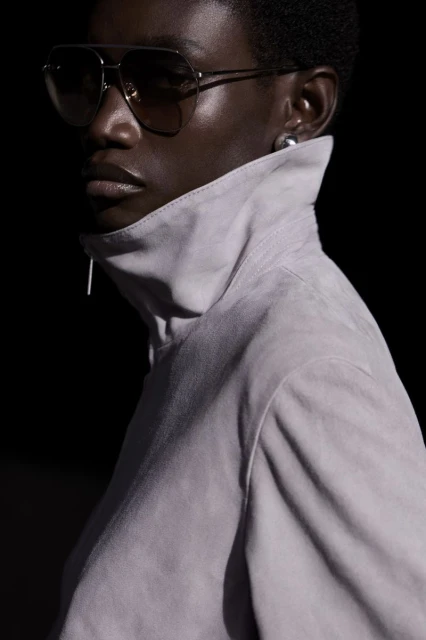 Close-up of a dark-skinned woman wearing aviator sunglasses and a high-collared jacket, against a dark background in studio lighting.