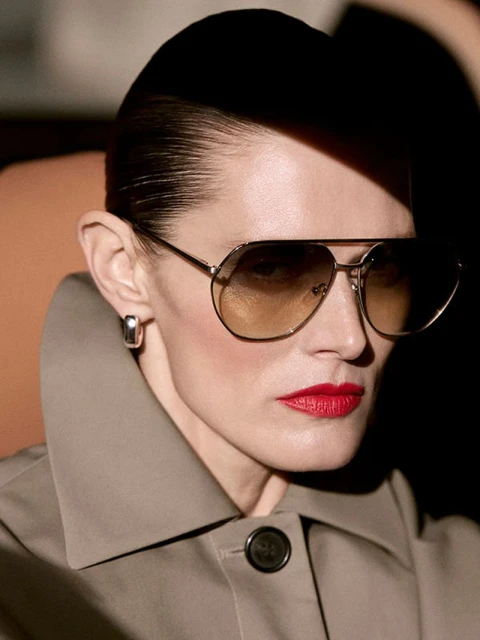 Close-up of an adult woman with sunglasses and red lipstick, wearing a tan coat with a button, against a partially dark background.