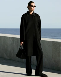 Model in black coat holding a handbag standing against a stone wall with ocean background