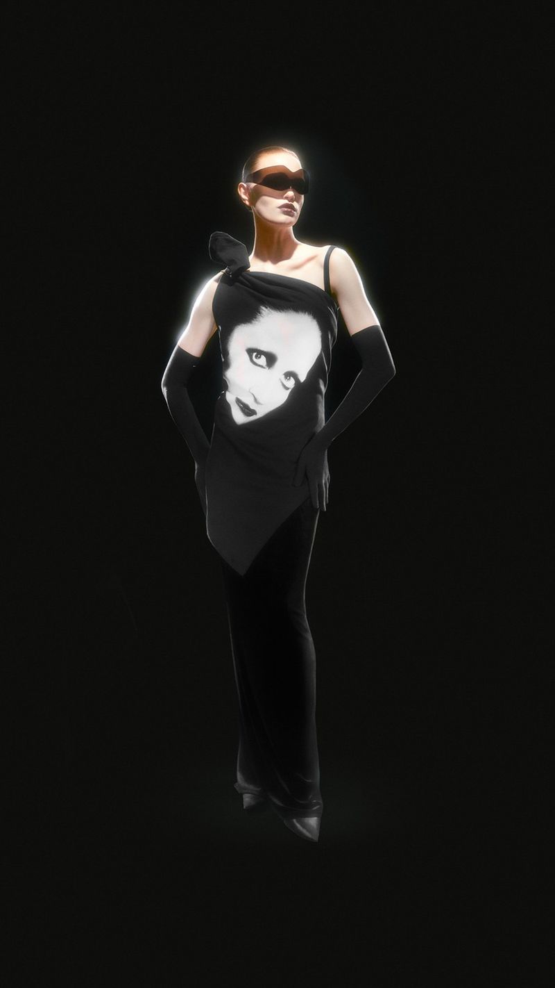 Fashion shot of woman in black dress with face graphic, wearing sunglasses and long gloves, against a black backdrop with soft lighting.