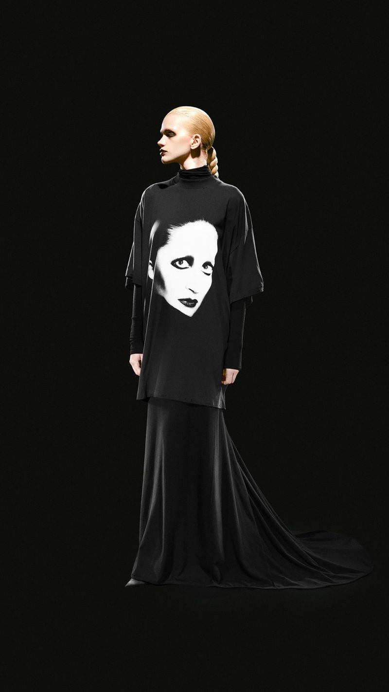 Fashion model stands in black floor-length gown with a portrait graphic on the front, against a black studio background in a full shot.