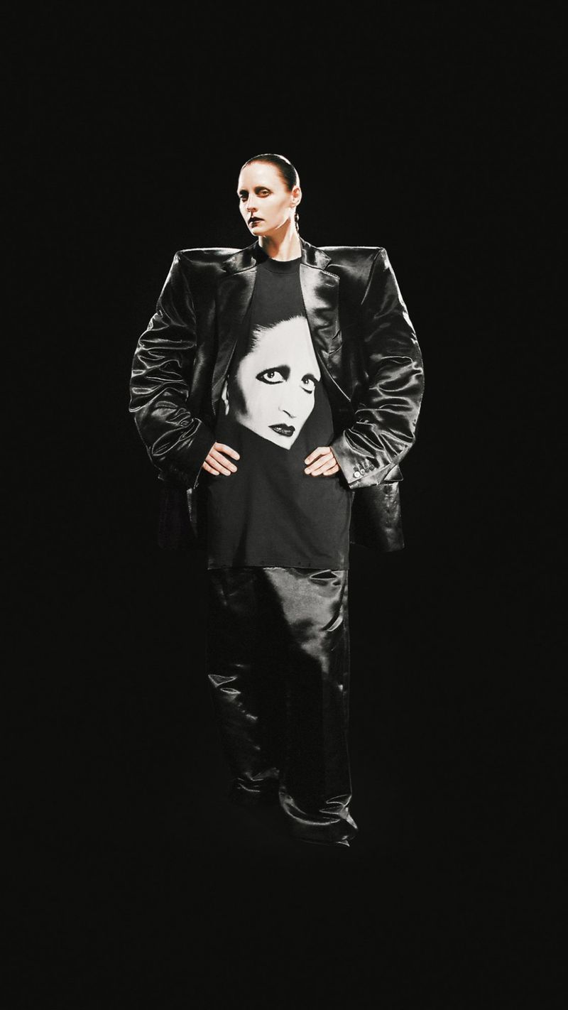 Adult female fashion model poses in a black jacket, black t-shirt with a face graphic, and black pants against a black backdrop.