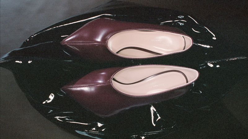 Two burgundy patent leather ballet shoes with pointed toes rest on a high-shine black surface, creating a sleek, reflective footwear product shot.