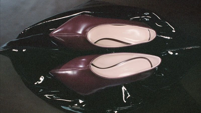 Two burgundy patent leather ballet shoes with pointed toes rest on a high-shine black surface, creating a sleek, reflective footwear product shot.