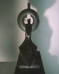 Black accessory sculpture displayed on a stand against a neutral white wall with strong directional shadow.