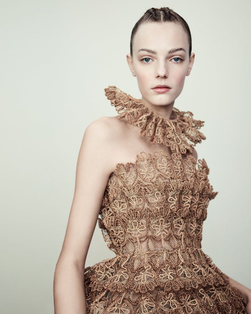 A woman in an elaborate, light brown lace dress with a ruffled collar poses against a neutral background, showcasing high fashion and intricate design.