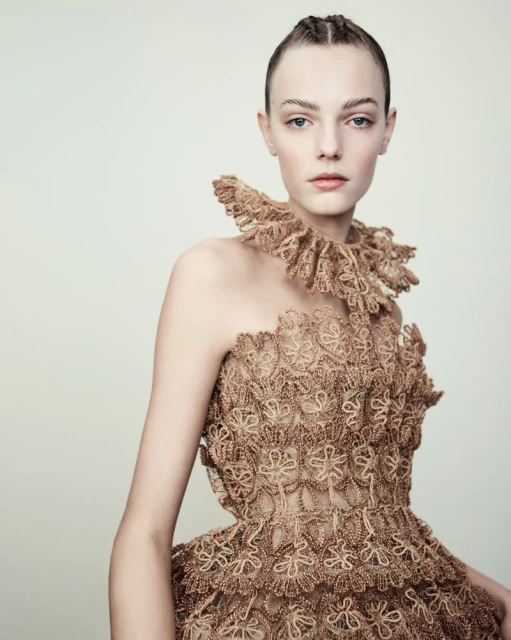 A woman in an elaborate, light brown lace dress with a ruffled collar poses against a neutral background, showcasing high fashion and intricate design.