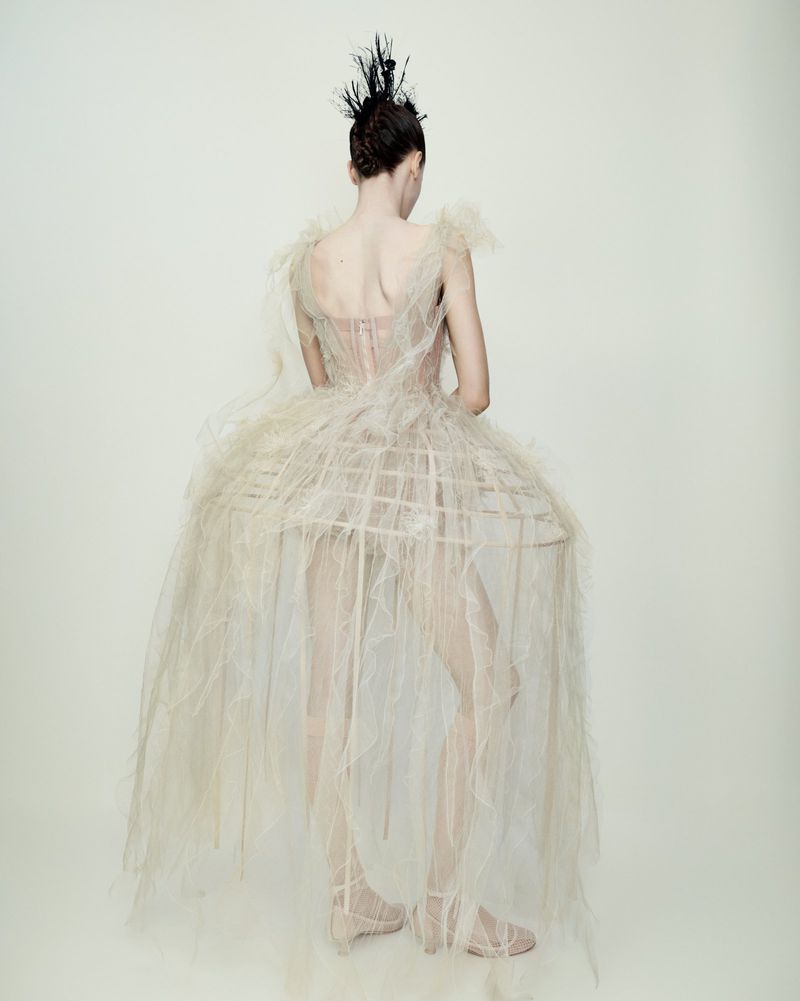 Back view of a fashion model posing in a tulle dress with a birdcage skirt and feathered hairpiece, set against a plain white backdrop.