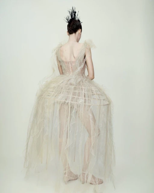 Back view of a fashion model posing in a tulle dress with a birdcage skirt and feathered hairpiece, set against a plain white backdrop.