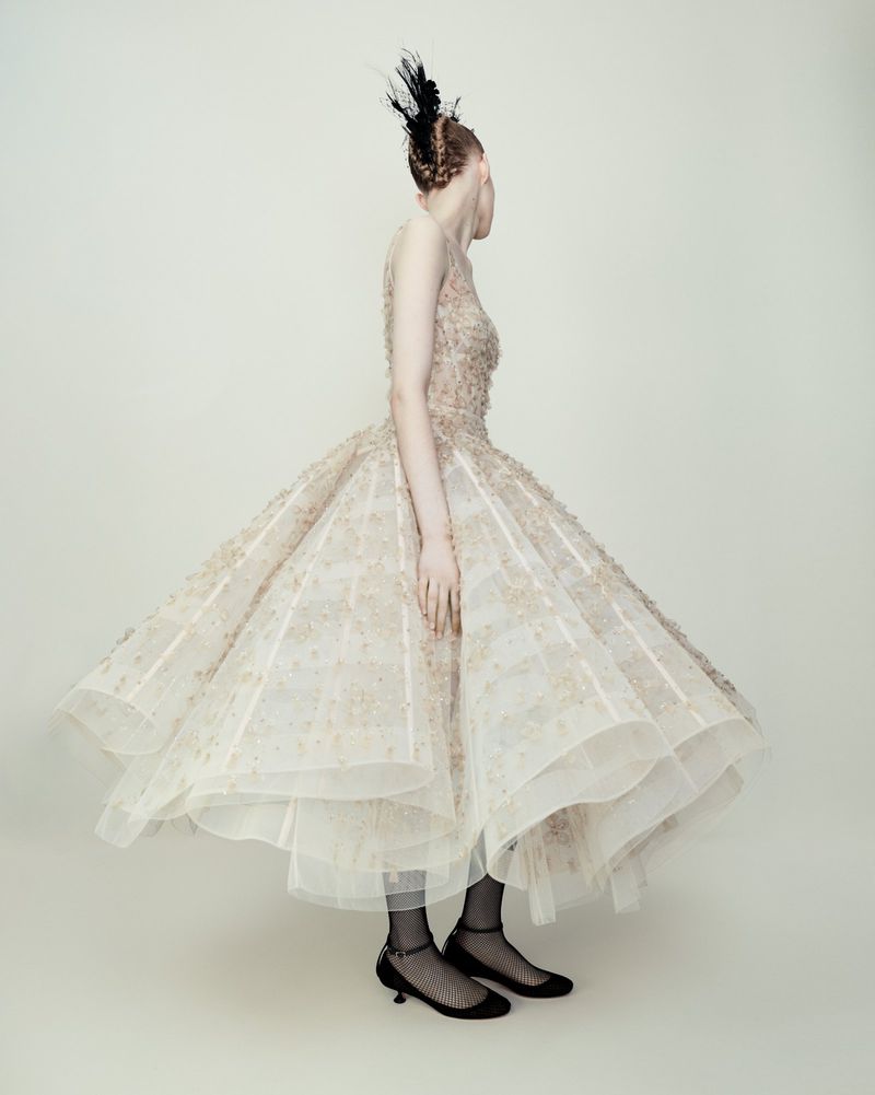 Woman in a formal gown with feather headpiece poses in a photo studio, back to the camera, showcasing fashion and elegant design.