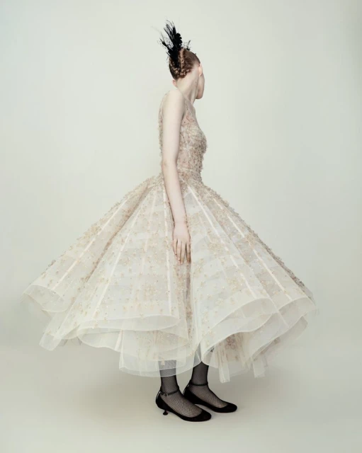 Woman in a formal gown with feather headpiece poses in a photo studio, back to the camera, showcasing fashion and elegant design.