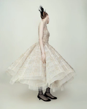 Woman in a formal gown with feather headpiece poses in a photo studio, back to the camera, showcasing fashion and elegant design.