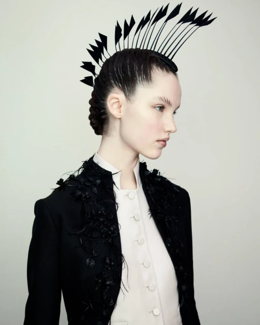 Side-view portrait of a slender adult woman with slicked-back dark hair, wearing a white high-neck buttoned shirt and black jacket with feather details.