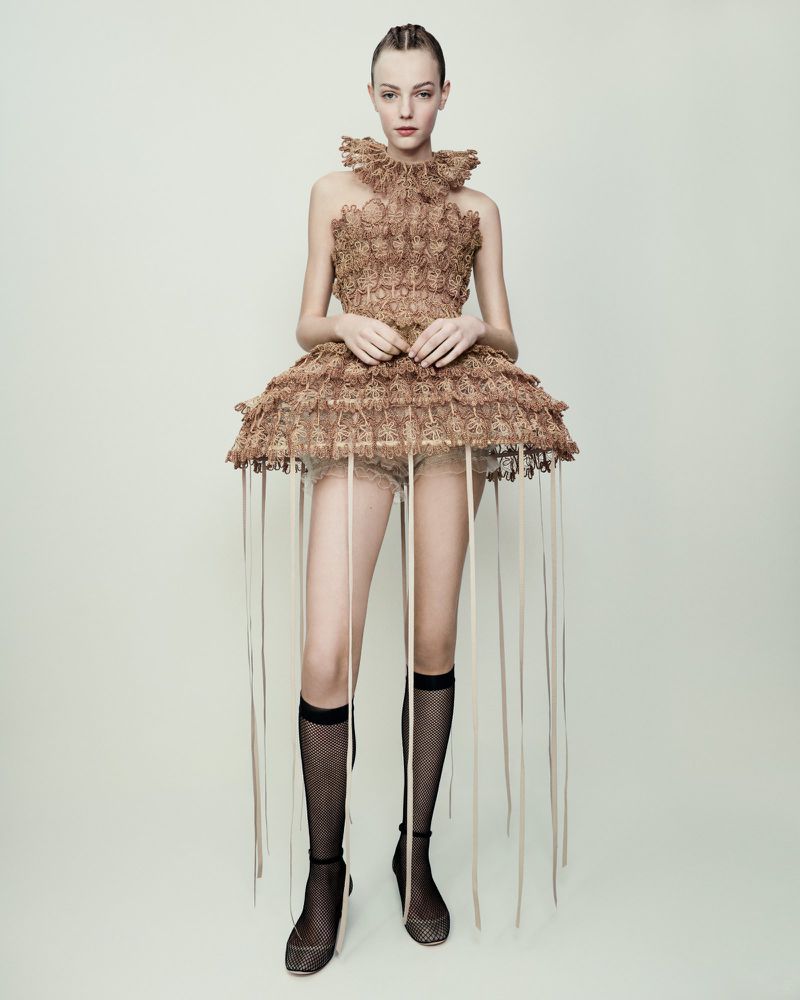 Fashion model in a sculptural beige dress with ribbons, standing against a light background in a studio, complemented by sheer black socks.