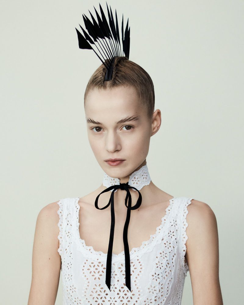 Close-up studio shot of a female fashion model with a decorative headpiece and white dress against a pale background.