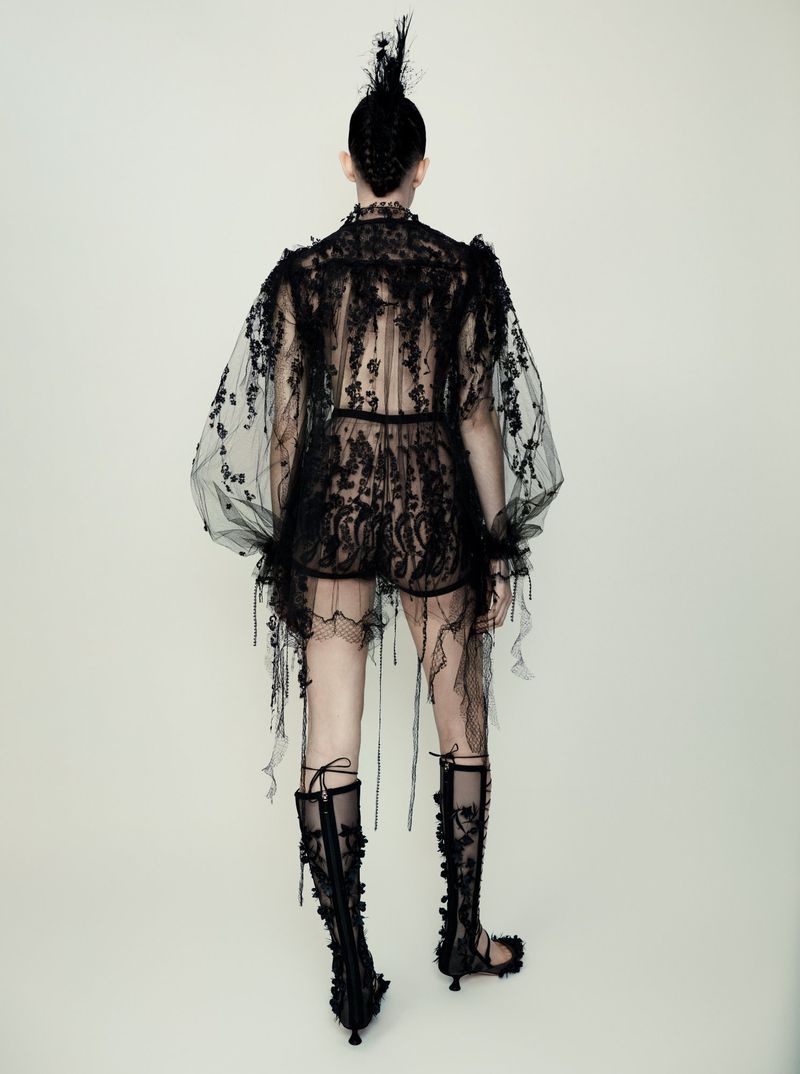 Back view of a model wearing a sheer black lace outfit with knee-high lace-up boots against a plain background in a fashion editorial.