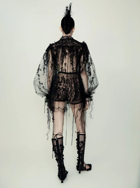 Back view of a model wearing a sheer black lace outfit with knee-high lace-up boots against a plain background in a fashion editorial.