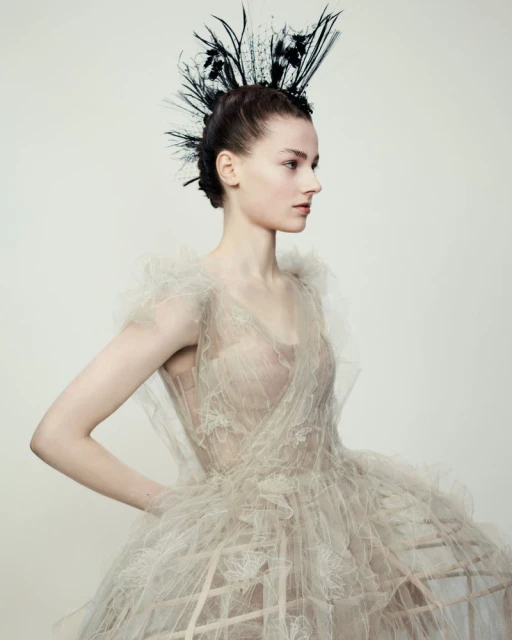 Elegant woman in ornate beige tulle dress and feathered headpiece poses in a studio portrait with a neutral light-grey background.