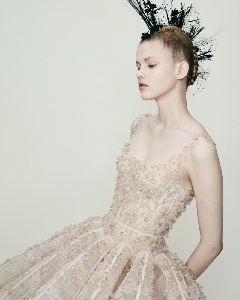 Woman poses in a blush-colored formal gown with floral appliques and dramatic feather headdress in a studio with soft, diffused lighting.