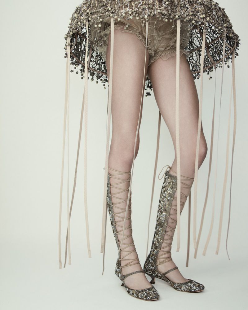 A woman is standing in a studio wearing a beaded dress and tall lace-up boots with ribbons hanging from the bottom of her dress.