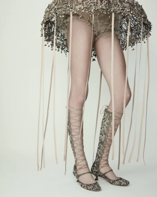 A woman is standing in a studio wearing a beaded dress and tall lace-up boots with ribbons hanging from the bottom of her dress.