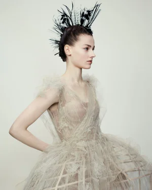 Model wearing a structured sheer gown and black feather headdress against a neutral background for Dior Haute Couture.