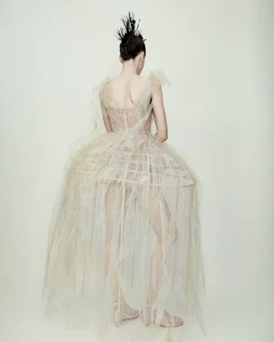 Model from behind wearing a sheer structured cage gown with voluminous tulle ruffles and a feathered headpiece against a plain backdrop.