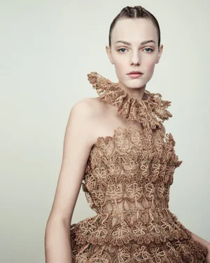 Model wearing an intricate brown lace haute couture dress with a ruffled high collar against a plain studio background.