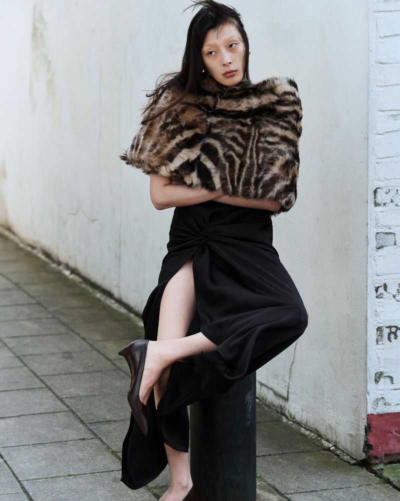 A woman in black dress poses in an outdoor setting with a furry shawl, one leg extended, showcasing a dark, pointed-toe heel.