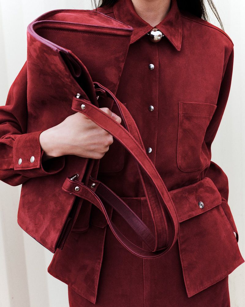 Woman in maroon suede jacket and skirt carrying a matching handbag. Studio shot with soft, diffused lighting and neutral background.