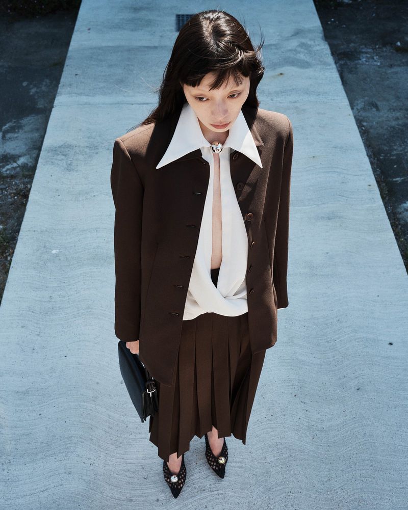 Model wearing brown jacket and white collared shirt standing on concrete path for Beaufille Resort 25