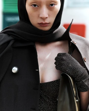 Fashion portrait of an adult woman with a black headscarf, coat with a silver button, gloves, and dress with black, white, and grey speckles.