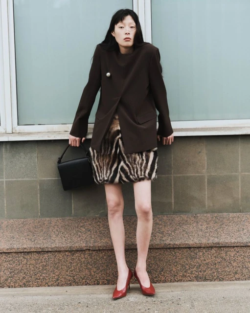 An Asian woman poses against a muted blue tile wall, wearing a dark brown blazer, patterned skirt, red heels, and carrying a black bag.