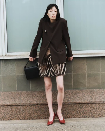 An Asian woman poses against a muted blue tile wall, wearing a dark brown blazer, patterned skirt, red heels, and carrying a black bag.
