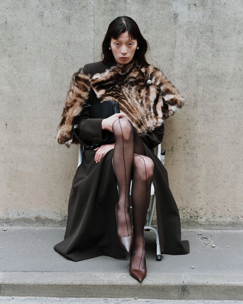 Elegant woman poses seated wearing fur stole, dark coat, sheer hose, and pumps against a concrete wall in an outdoor urban environment.